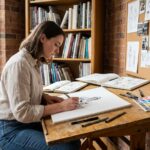Jeune femme autodidacte dessinant des illustrations dans son studio à domicile, entourée de carnets de croquis et de références artistiques